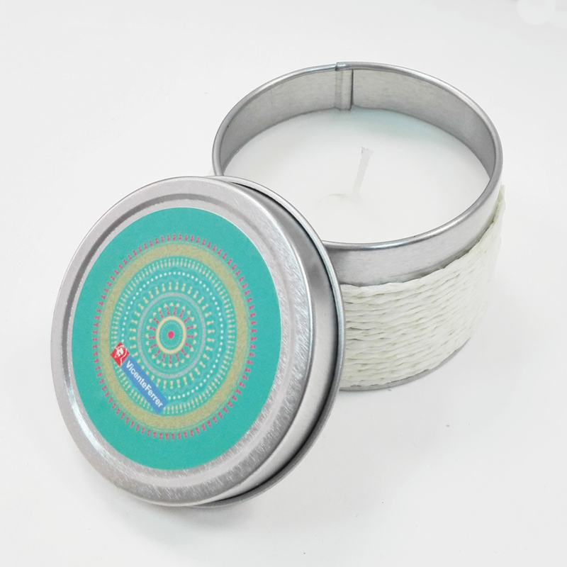 Vela perfumada de vainilla en recipiente metálico plateado  y tapa decorativa colocada al frente mostrando su diseño circular